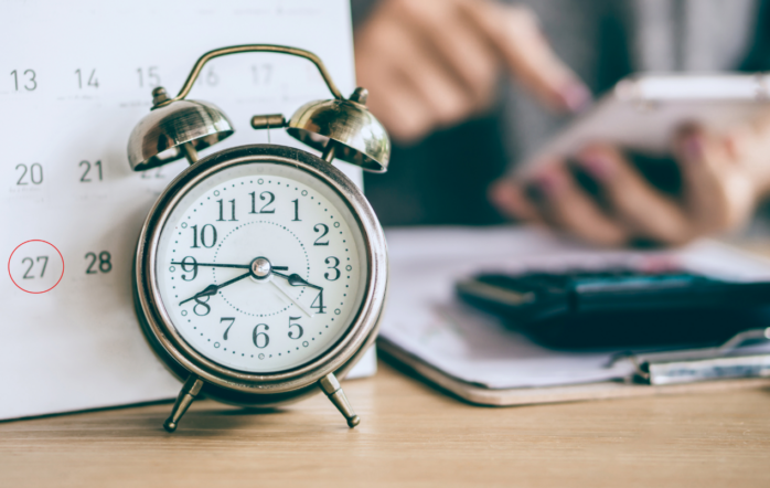 Clock and calendar in the foreground with a businessman working in the background. Concept of time's significance in business and proper time to sell business
